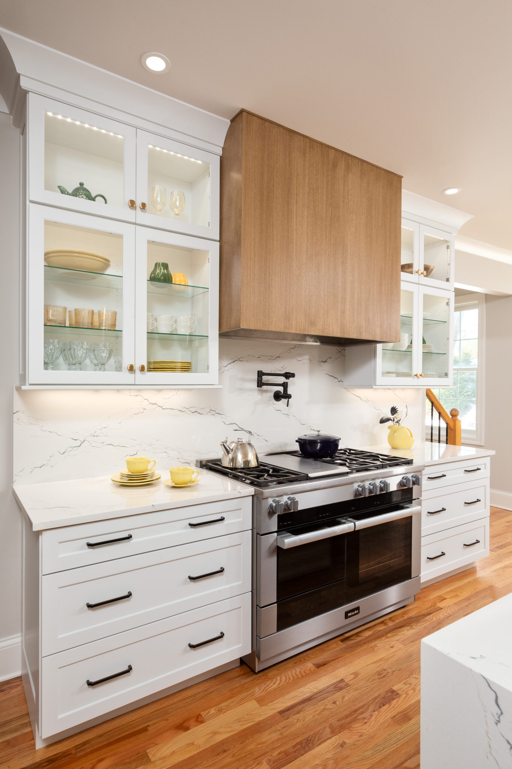 Modern Niskayuna kitchen with white shaker cabinets, quartz backsplash, glass upper cabinets, and custom rift-cut oak range hood above stainless steel gas range.